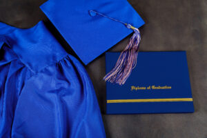 blue graduation cap and gown laying on gray background with blue diploma folder that says diploma of graduation in gold letters