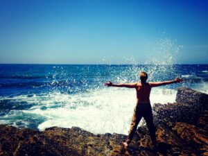 white man with cargo pants stands facing the ocean wave with his arms stretched